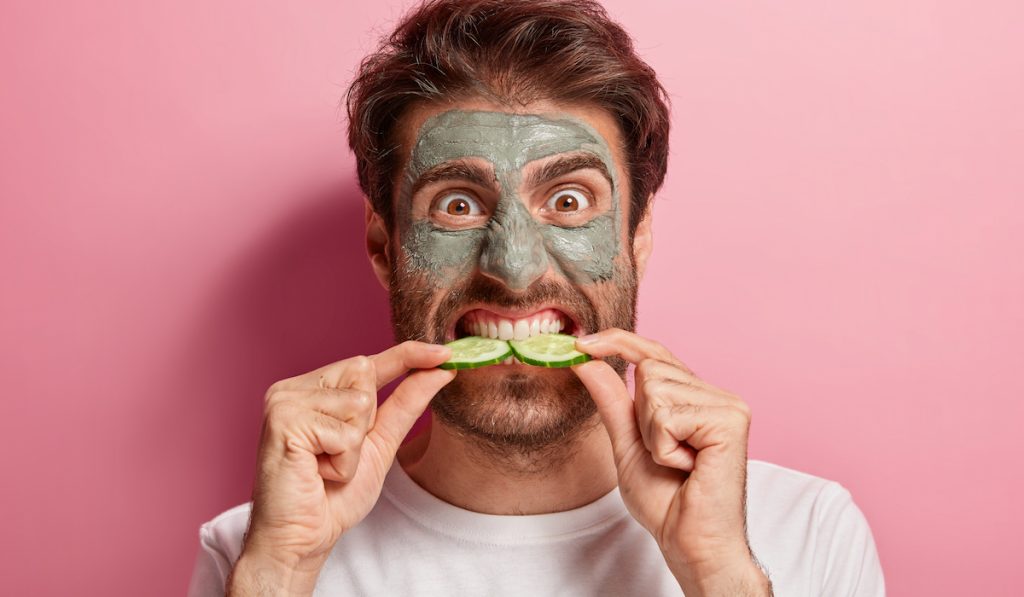man with mud mask biting two slices cucumber