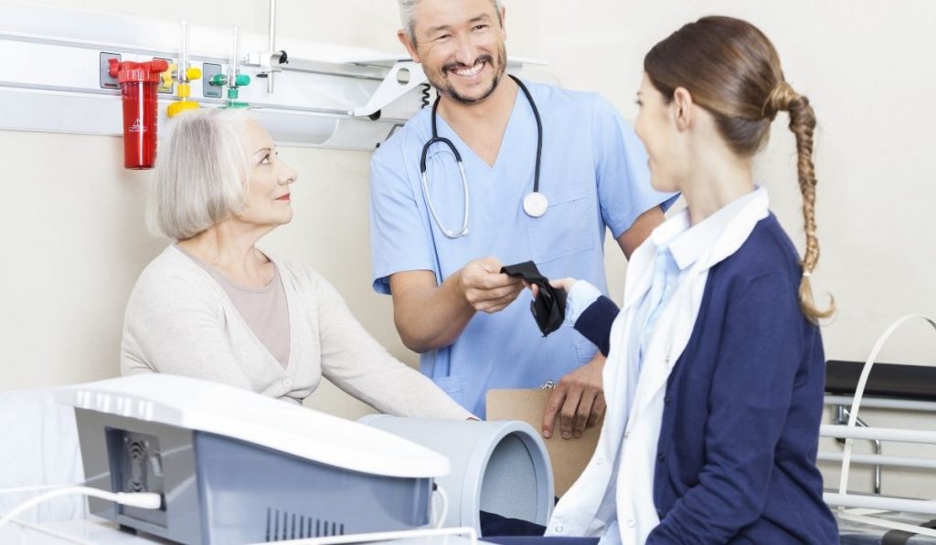 physiotherapist giving belt to nurse while senior patient is getting ready for her electromagnetic therapy - ss220324